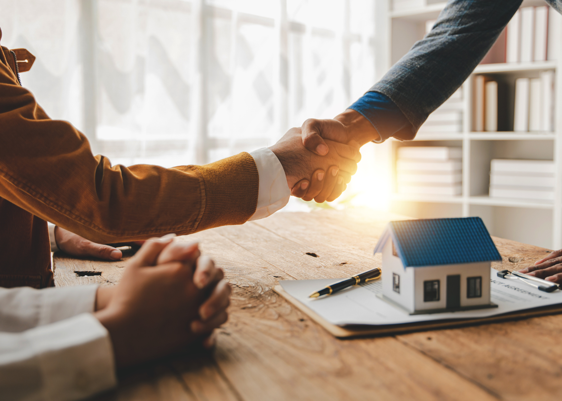 Two people shaking hands with contract over a desk next to another person with paperwork on the desk